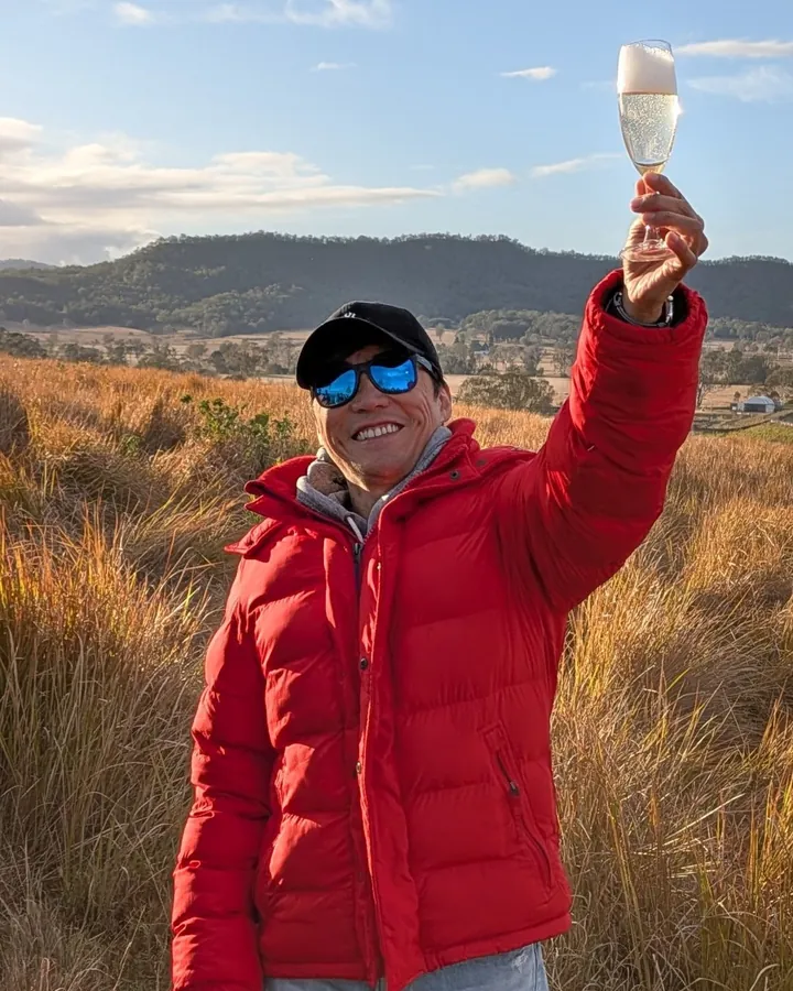 Robin Chien in a red jacket, raising a glass in a field of golden grass